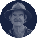 Smiling elderly man wearing a wide-brimmed hat and a checkered shirt against a dark circular background.