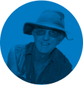 Portrait of a smiling person wearing glasses, a wide-brimmed hat, and a jacket against a blue circular background.