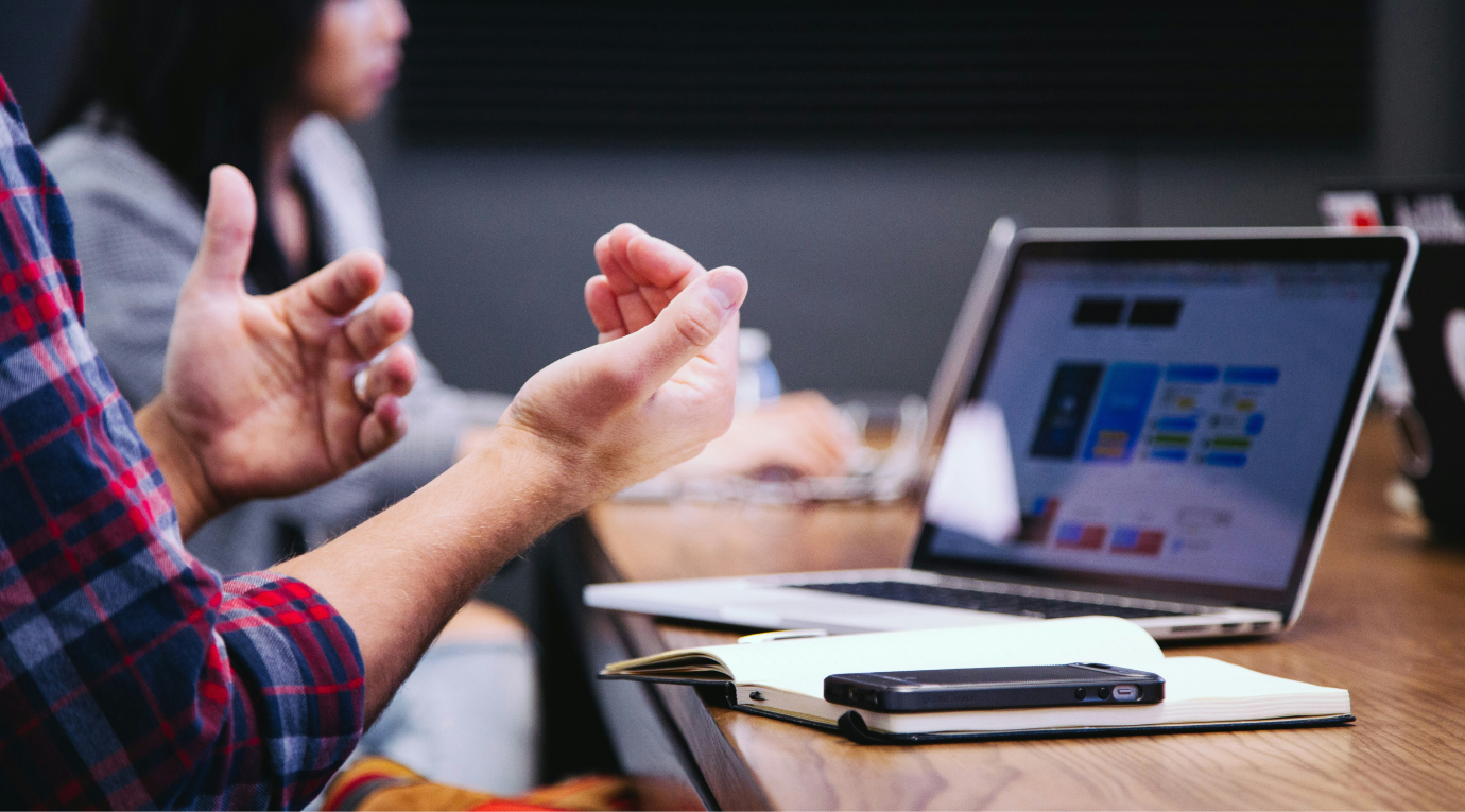 Person gesturing with hands in a meeting with a laptop, notebook, and phone on the table.
